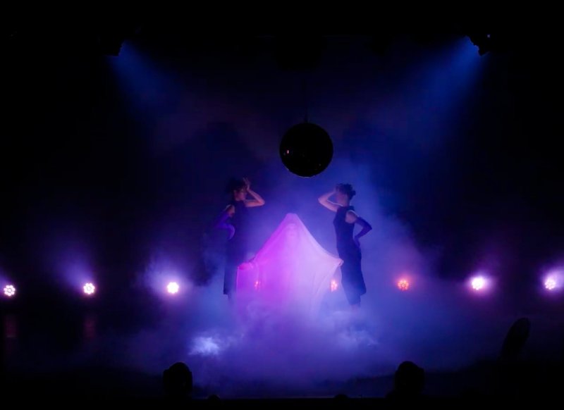 Two figures in dark clothing stand on a stage amidst purple and blue fog and spotlights, with a disco ball overhead.