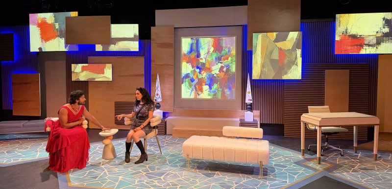 Two women on a stage set with abstract art, blue lights, and geometric patterns. One in a red dress, one in a sparkly dress.