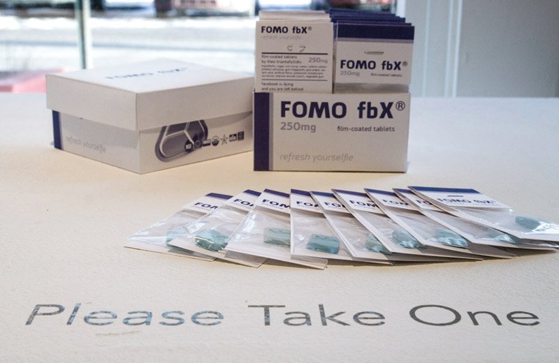 White boxes of "FOMO fbX" tablets and small clear packets of blue pills on a white table with "Please Take One" printed on it.