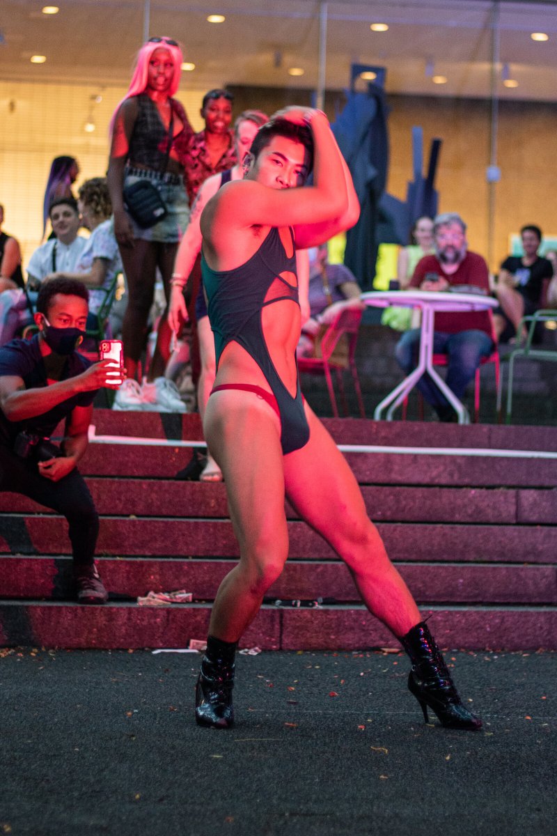 A person in a dark teal bodysuit and black high-heeled boots poses on a city street at night, with spectators behind.