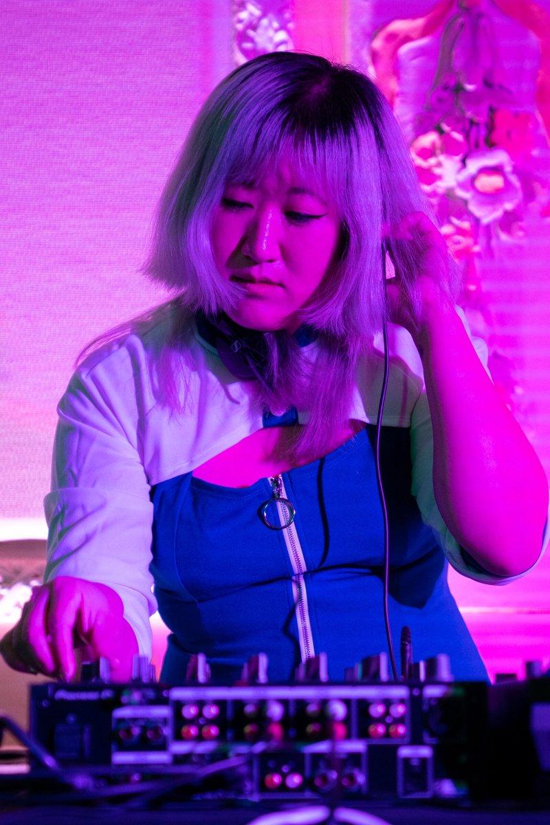 Female DJ with silver hair and blue dress, adjusting headphones while mixing on a console under bright pink light.