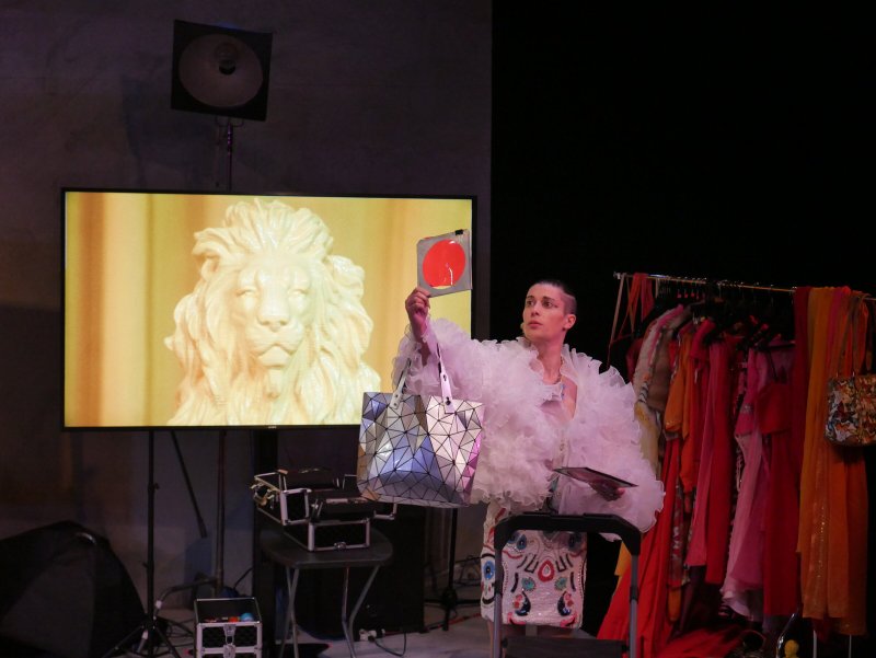 Performance art with a person in a ruffled white top holding a silver geometric bag and a red circle, next to a screen displa