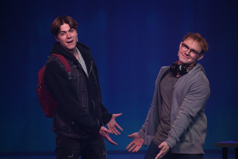 Two young men, one with a red backpack, the other with headphones, pose with open hands and smiles against a blue-lit stage.