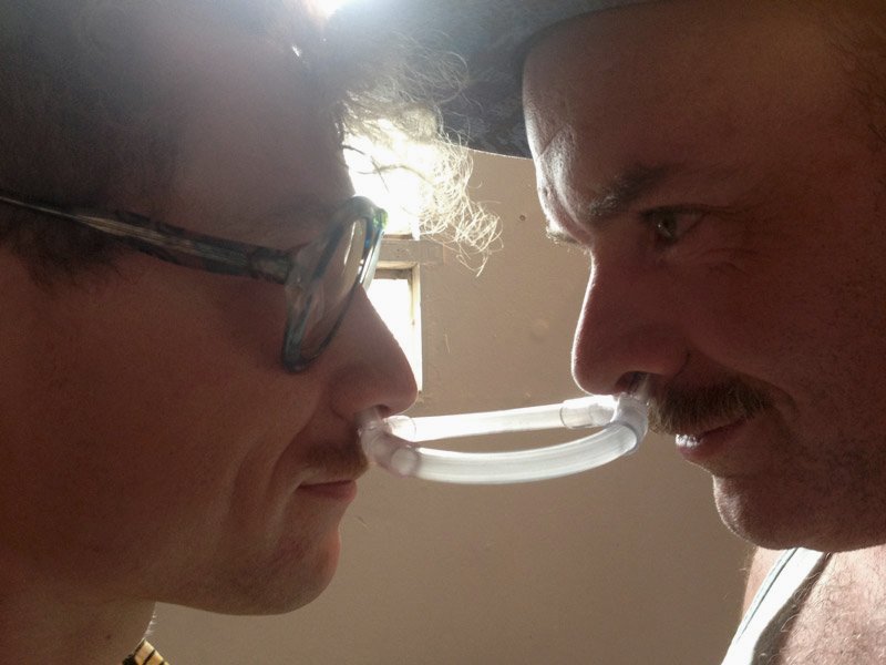 Two men face each other, connected by a clear plastic tube through their nostrils. One wears glasses, the other a hat.