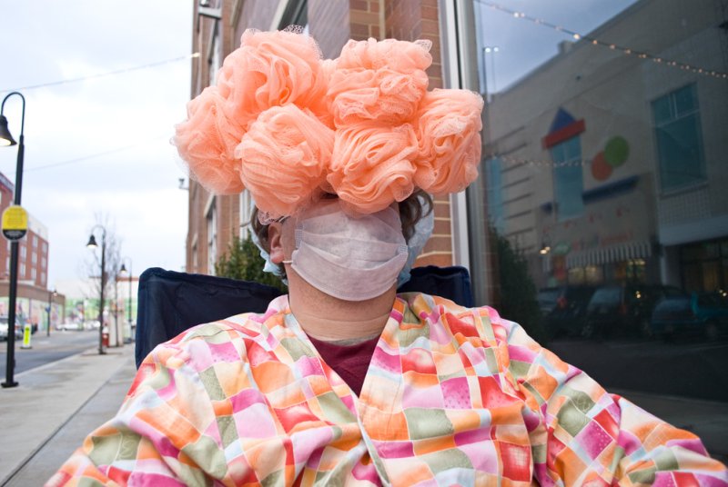 Person in colorful robe and surgical mask, with a large, fluffy peach-colored headpiece, sitting outdoors.