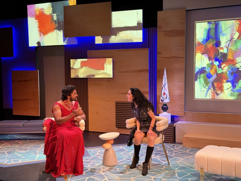 Two actors on stage with abstract art projections. Man in red dress gestures, woman in sequined dress sits opposite.