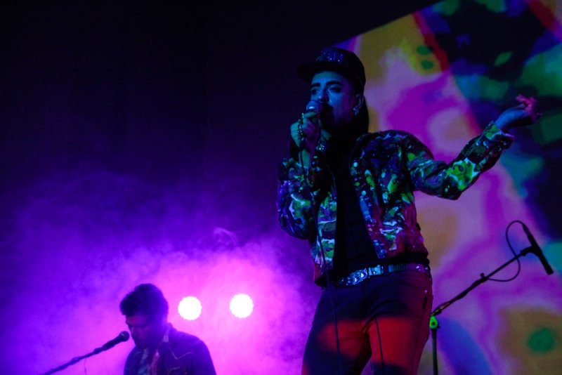 A singer in a colorful jacket and cap performs under purple and blue stage lights with a blurred psychedelic background.