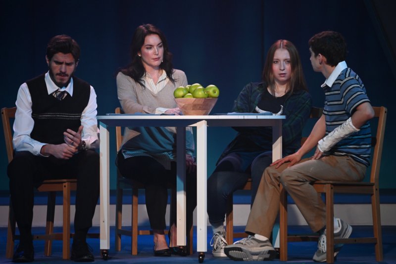 Four actors on stage, two men and two women, seated around a table with a bowl of green apples.