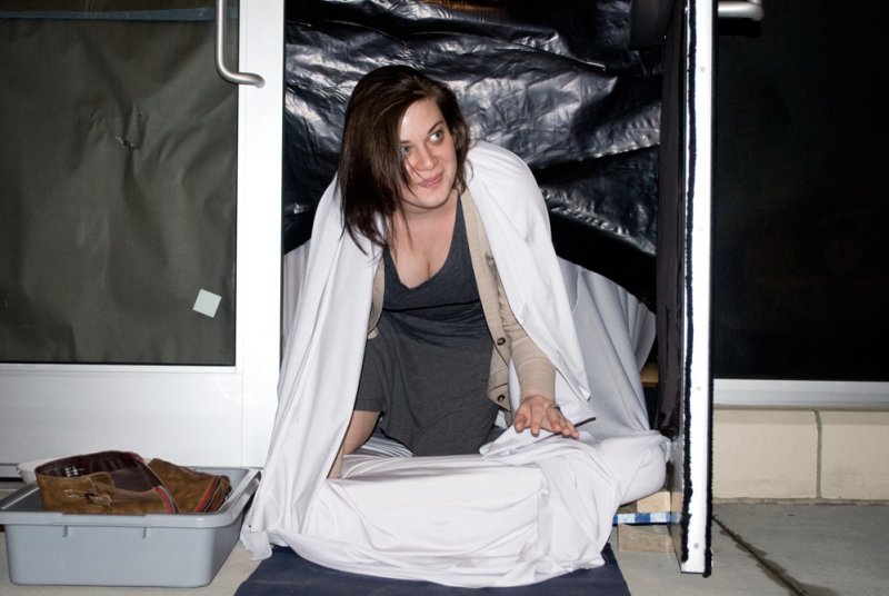 Woman with dark hair, wrapped in white fabric, peeking out from a dark, tarp-covered enclosure.
