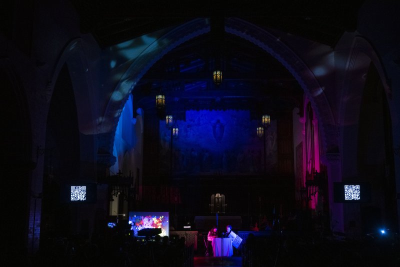 Dark church interior with blue and pink lights illuminating arches, a mural, and two performers at a console with a colorful screen.