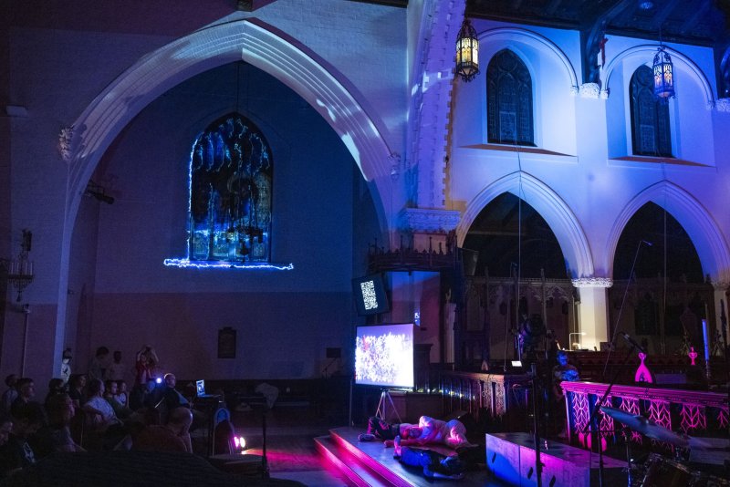 Scott N Andrew's "Vox Humana" in a church, with purple and blue lights illuminating a screen, instruments, and an audience.