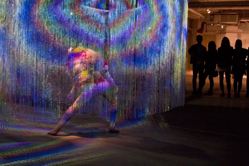 Performance art with a dancer in colorful costume against a shimmering, iridescent curtain, casting rainbow light.
