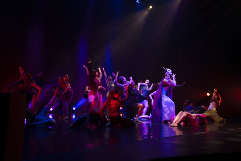 Dancers perform on stage in colorful costumes, lit with blue and purple light. One dancer wears a long gown and cape.