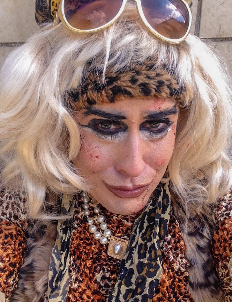 Close-up of a person in drag with blonde wig, leopard print, pearl necklace, heavy eye makeup, and sunglasses on head.
