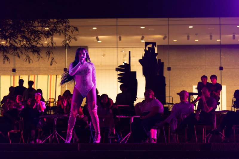 A performance scene bathed in purple light. A singer in a shimmering bodysuit and thigh-high boots stands on stage, microphone in hand, facing an audience seated in rows. A modern building with large windows forms the backdrop.