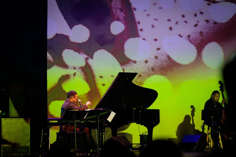 Man in dark suit playing a grand piano on stage, with a bassist to his right, against a vibrant yellow and purple abstract projection.