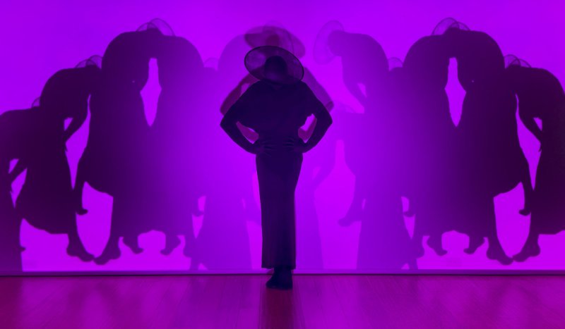A person in a wide-brimmed hat and long dress stands silhouetted against a vibrant purple wall, casting a large shadow.