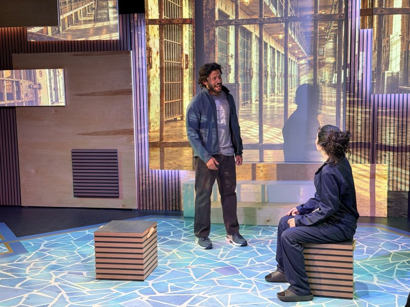 A man stands center stage, smiling at a seated woman, both in casual clothes, against a backdrop of projected prison cells.