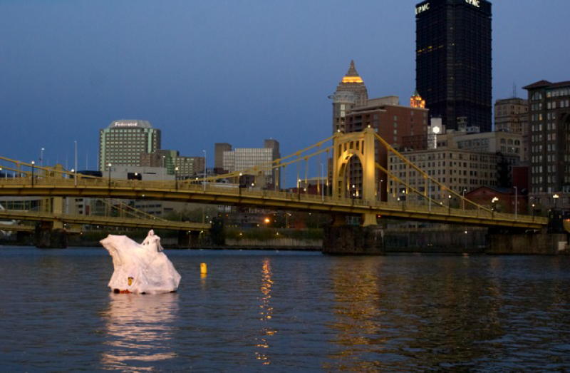 White sculpture of a figure on a raft in a river, with a yellow bridge and city skyline at dusk.