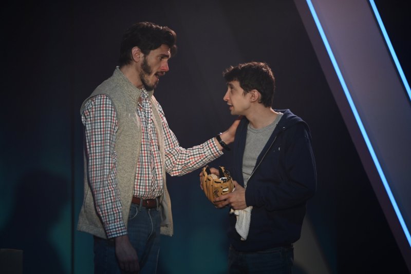 Two men on a dark stage with blue light. One, in a plaid shirt and vest, holds the other's shoulder. The second man, in a hoodie, holds a baseball glove.