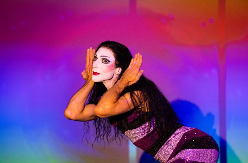 Performer in drag with white face paint, red lips, and dark eye makeup, wearing a sparkly purple and black striped dress, hands framing face, against a vibrant pink, purple, and bl