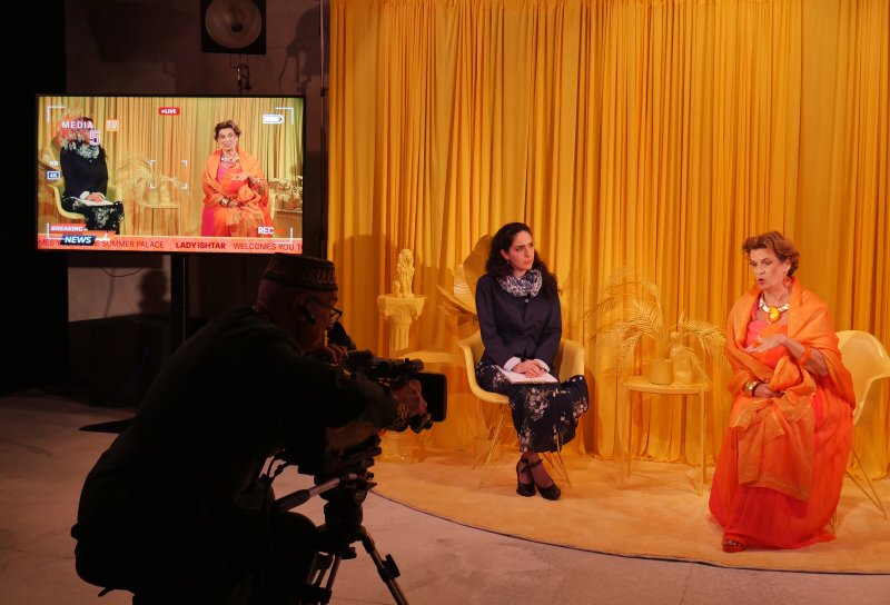 A man films two women on a yellow set, one in orange, the other in a patterned dress, with a monitor displaying the live feed