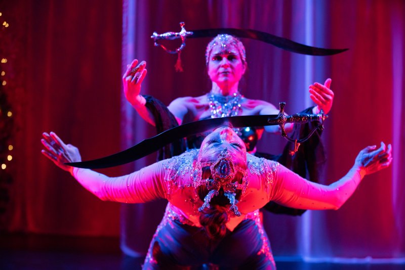 Two women in bejeweled costumes balance swords on their heads and hands, bathed in dramatic red and purple stage lighting.