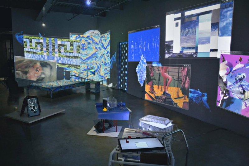 Gallery installation with projected digital images on walls, displaying figures, patterns, and landscapes in blue, purple, and green hues. Foreground includes blue furniture and tech equipment.