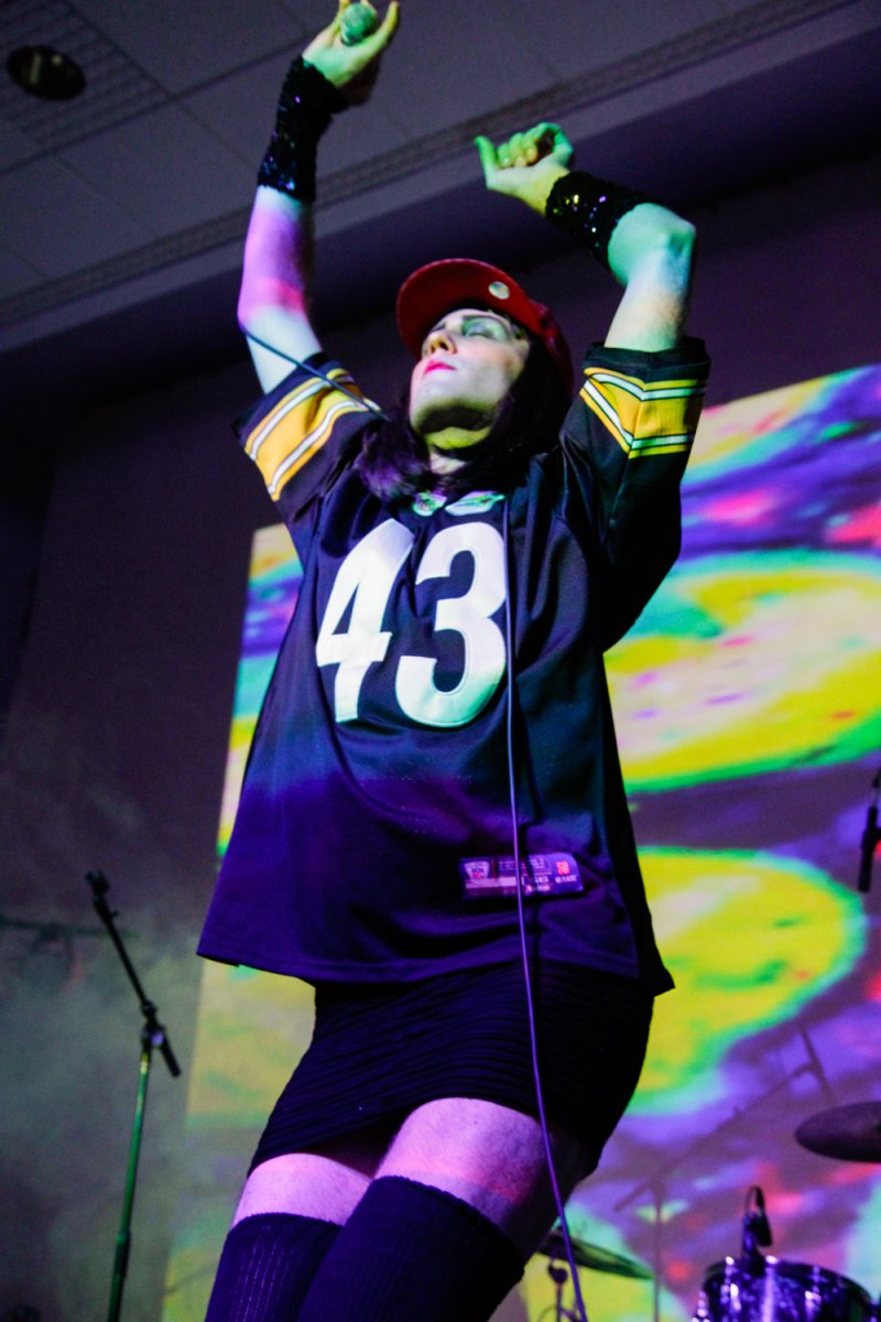Performer in jersey, red cap, and black skirt raises arms on stage with colorful background.
