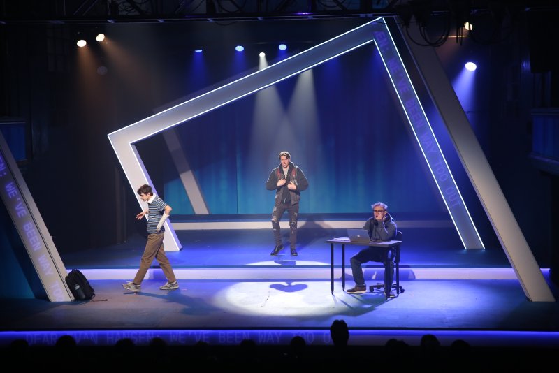 Three men on a dark stage with blue lighting. One walks left, one stands center, one sits at a desk right. A large white geometric frame surrounds them.