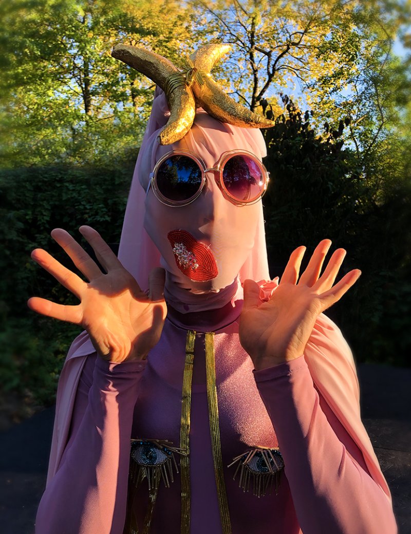 Person in pink bodysuit, gold banana on head, round sunglasses, red beaded lips, hands raised, outdoors.