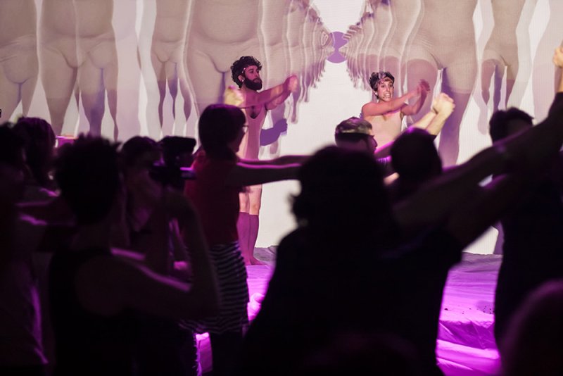 Two performers in nude bodysuits on stage, arms outstretched, in front of a projection of repeating nude figures.