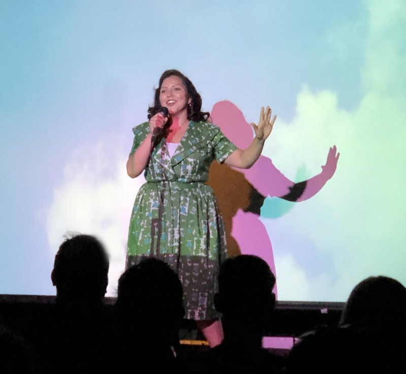 A woman in a green patterned dress speaks into a microphone on stage. Behind her, a light blue and green backdrop with a pink shadow of her. Silhouetted heads of the audience are visible in the foreground.