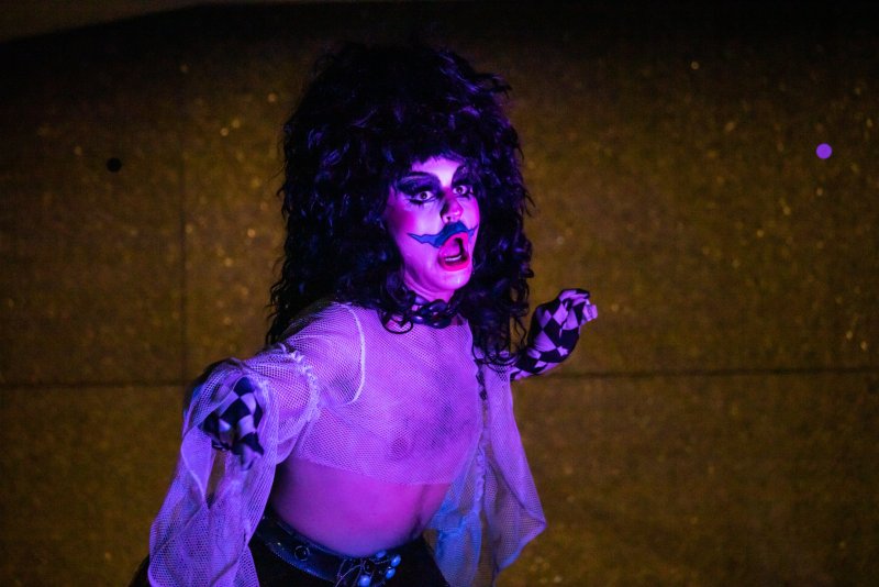Performer with dark, teased hair and theatrical makeup, including a blue painted mustache, in a purple-lit scene. They wear a sheer top with checkered sleeves and a black belt.