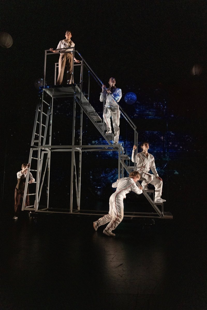 Five performers in white and brown costumes interact on a multi-level metal structure against a dark, starry background.