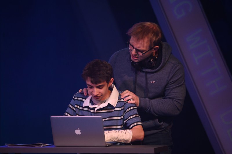 Two young men on a dark stage, one with a cast on his arm, looking at a laptop, the other standing behind him with hands on his shoulders.