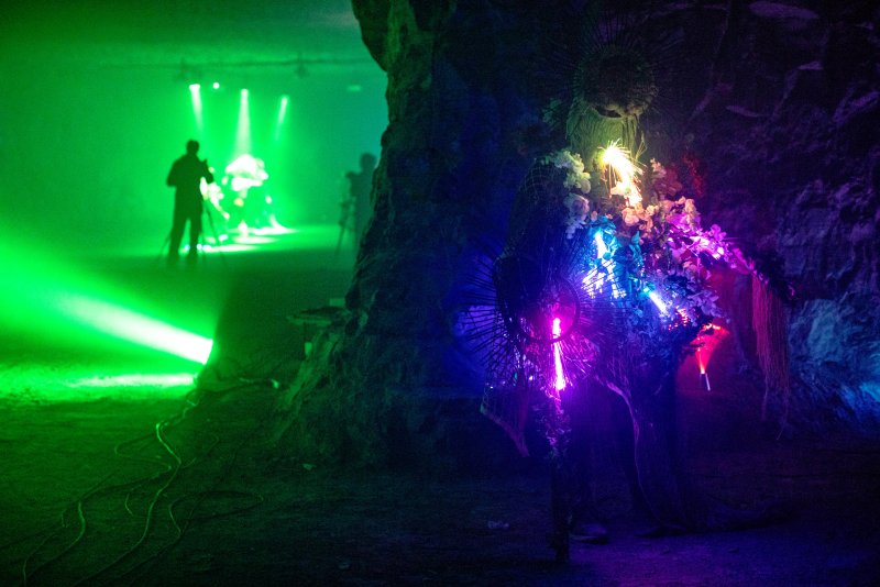 Dark cave interior with glowing purple and blue light on the right, and a green-lit figure in the distance.
