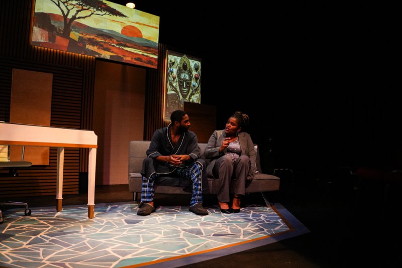 Two actors on a stage set with a savanna painting and a tribal mask, a man in a robe and a woman in a suit.