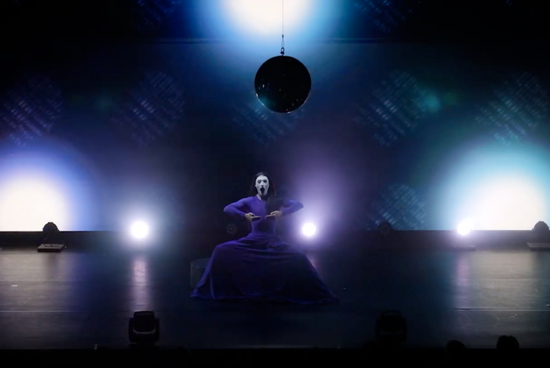 Performer in a long purple dress and white face paint, squatting on a dark stage under a disco ball and bright blue and white lights.