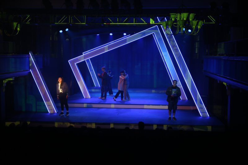 Stage production with actors under blue lights, framed by glowing white geometric structures.
