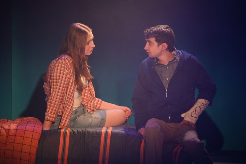 Two young actors on a stage with blue lighting. A woman in a plaid shirt sits left, a man in a blue hoodie with a cast sits right.