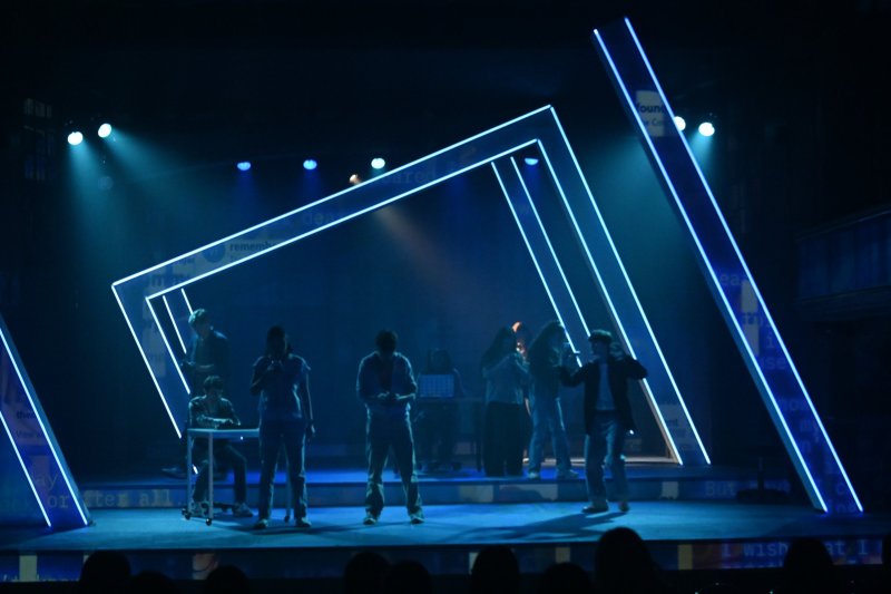 Stage production with actors in dark clothing under blue lights, framed by glowing geometric lines.