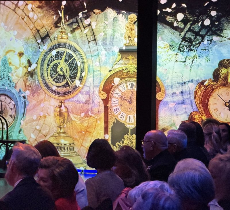 Audience watches a vibrant projection of ornate, antique clocks in gold, teal, and brown, with falling white petals on a textured background.