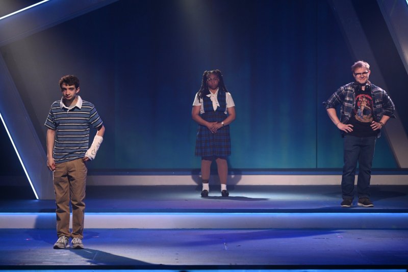 Three actors on a stage with blue lighting; a young man in a striped shirt, a young woman in a plaid dress, and a man in a plaid shirt.