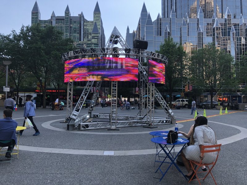 Outdoor video installation: metal scaffolding supports a screen displaying vibrant red, purple, and yellow abstract patterns.