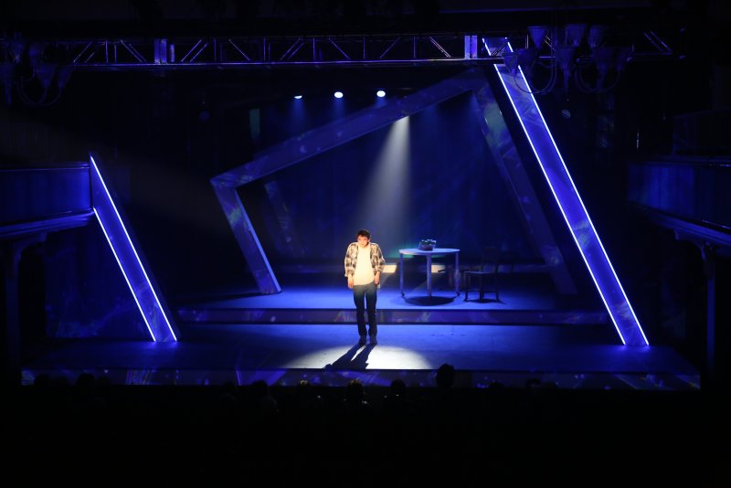 A lone figure stands center stage under a spotlight, surrounded by geometric structures with glowing blue edges against a dark background.