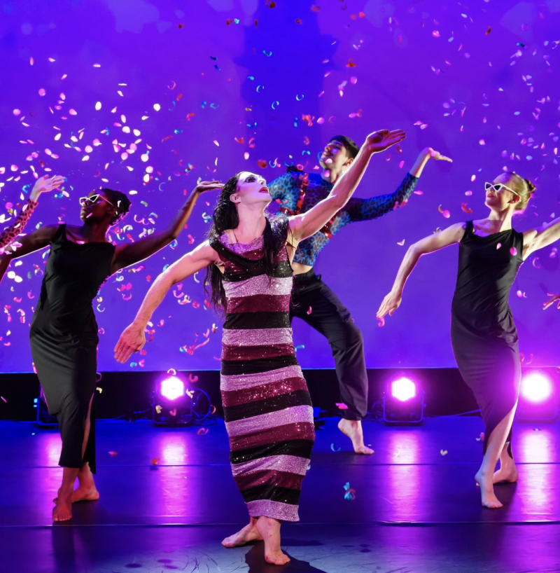 Four dancers on a purple stage with falling confetti. Center dancer wears a striped gown, others wear black dresses or patter