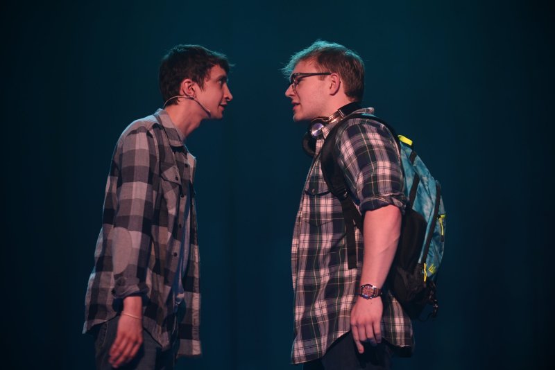 Two young men in plaid shirts face each other, one with a headset, the other with glasses and a backpack, against a dark background.