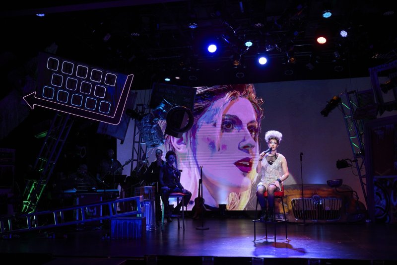 Stage performance with a singer in a white wig, fishnets, and a sequined top, seated before a large screen displaying a stylized face.