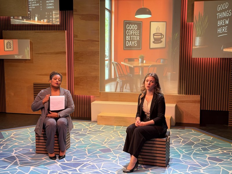 Two women sit on striped boxes on a blue geometric floor, one holding papers, against a backdrop of cafe scenes and menus.